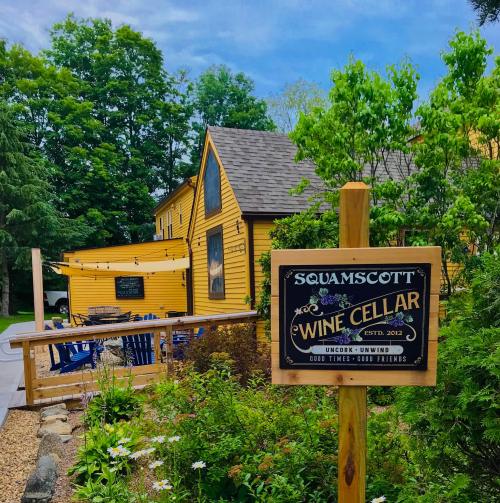 A charming yellow house with a sign reading "Squamscott Wine Cellar" surrounded by greenery and outdoor seating.