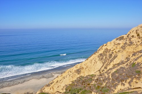 A serene coastal view featuring sandy cliffs, gentle waves, and a clear blue sky over the ocean.