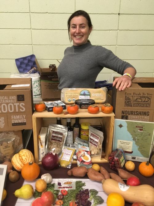 A smiling woman stands behind a table filled with fresh produce and local food products.