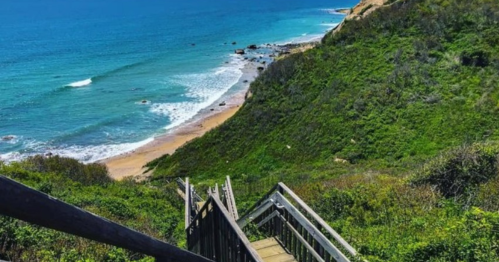 A wooden staircase leads down to a sandy beach surrounded by lush green hills and a blue ocean.