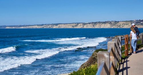 A couple stands by a wooden railing, overlooking waves crashing on rocky shores under a clear blue sky.