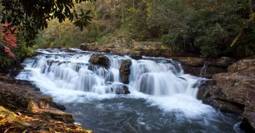 A serene waterfall cascades over rocky ledges, surrounded by lush greenery and autumn foliage.
