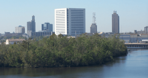 City skyline with modern buildings and greenery along a riverbank under a clear blue sky.