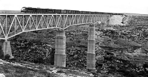 A vintage train crosses a high bridge over a rocky canyon, with freight cars visible in the background.