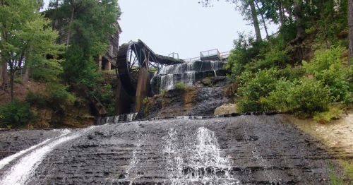 A scenic view of a watermill with a large wheel, cascading water, and lush greenery surrounding the rocky terrain.