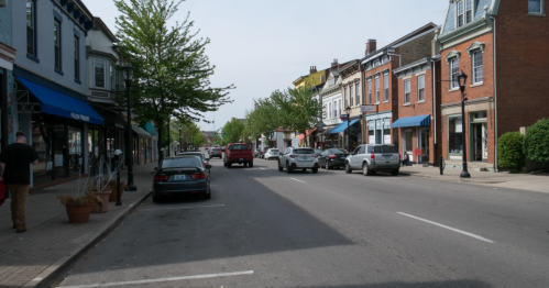 A quiet street lined with shops and trees, featuring parked cars and a few pedestrians on a sunny day.