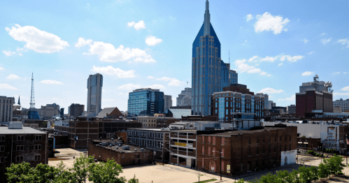 A city skyline featuring modern buildings and a prominent tower under a blue sky with scattered clouds.