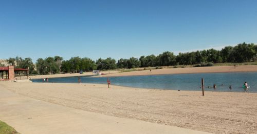 A sandy beach area with a calm water body, trees in the background, and people enjoying the water on a sunny day.