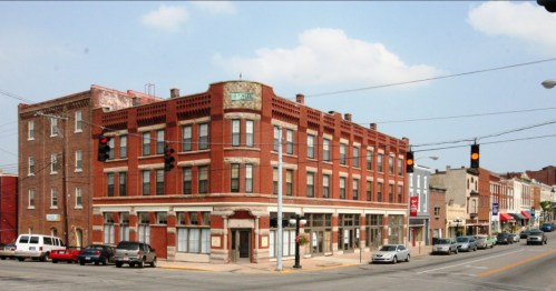 Historic brick building at a street intersection, with parked cars and shops visible in the background under a blue sky.