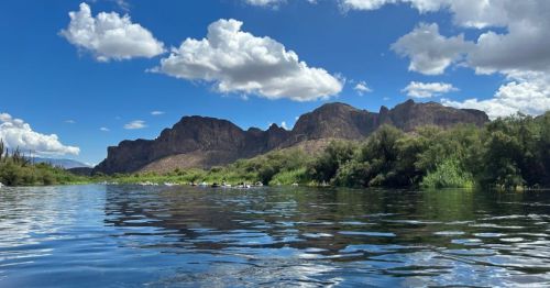 A serene river scene with mountains in the background and fluffy clouds in a blue sky. Lush greenery lines the water's edge.