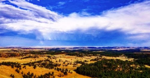 A vast landscape featuring rolling hills, green trees, and a bright blue sky with scattered clouds.