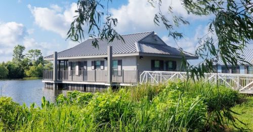 A modern house on stilts by a calm lake, surrounded by lush greenery and blue skies with fluffy clouds.