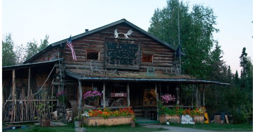 A rustic log cabin store with an "Open" sign, surrounded by flowers and American flags, set in a natural landscape.