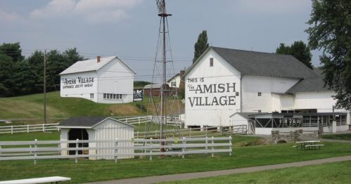 A rural scene featuring white buildings with signs for "Amish Village" and "Jewish Village," surrounded by green fields.