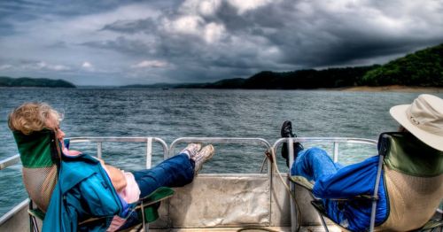 Two people relax in chairs on a boat, facing the water under a cloudy sky.