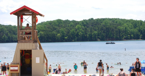 A lifeguard tower on a beach with people swimming and relaxing, surrounded by trees and a lake in the background.