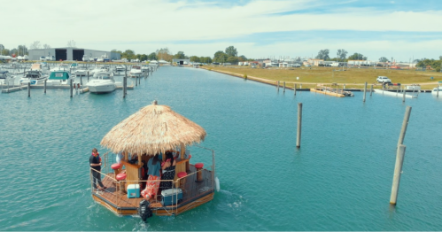 A tiki-style boat with people on it floats in a marina surrounded by boats and a clear blue sky.