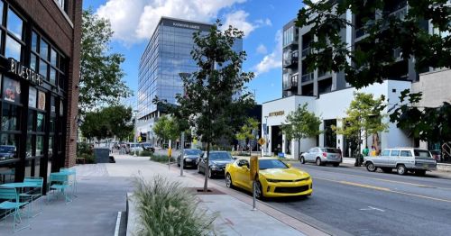 A city street lined with trees, parked cars, and modern buildings under a blue sky with clouds.