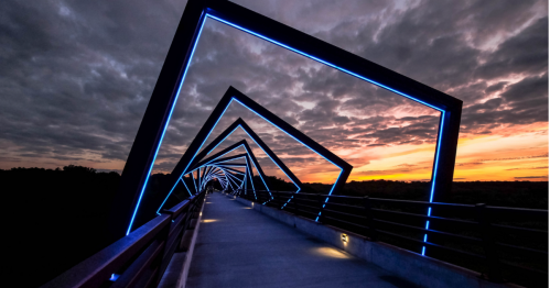 A modern bridge with illuminated geometric frames against a dramatic sunset and cloudy sky.