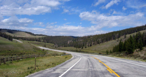 A winding road through green hills and trees under a blue sky with scattered clouds.