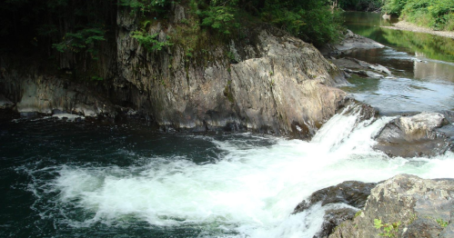 A serene river flows over rocky terrain, surrounded by lush greenery and calm waters.