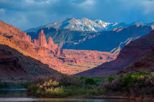 A scenic view of red rock formations and snow-capped mountains under a cloudy sky, with a river in the foreground.