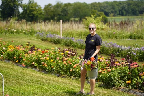 A person in sunglasses holds a bucket of flowers, standing in a vibrant garden filled with colorful blooms.