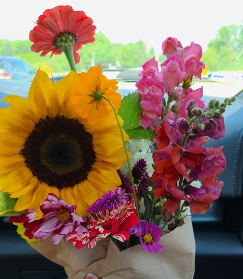 A vibrant bouquet featuring a sunflower, snapdragons, and various colorful flowers, held inside a car.