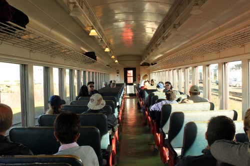Interior of a vintage train car with passengers seated on either side, sunlight streaming through the windows.