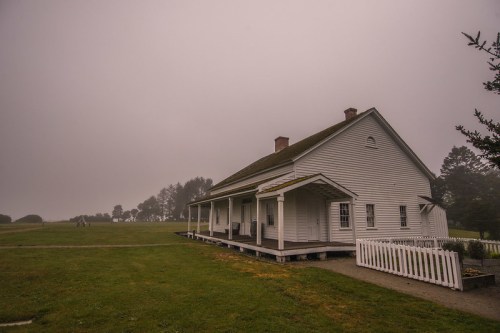 A white, two-story house surrounded by fog, with a grassy area and trees in the background.