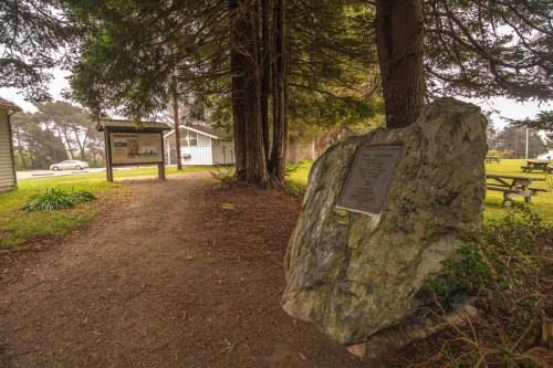 A stone monument with a plaque beside a path leading to a building, surrounded by trees and grassy areas.