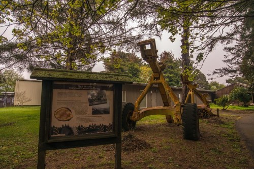 A yellow piece of machinery beside an informational sign in a grassy area, surrounded by trees and a building in the background.