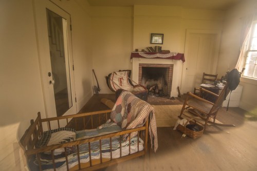 A cozy, rustic room with a fireplace, rocking chairs, and a wooden crib draped in a quilt. Natural light filters in.