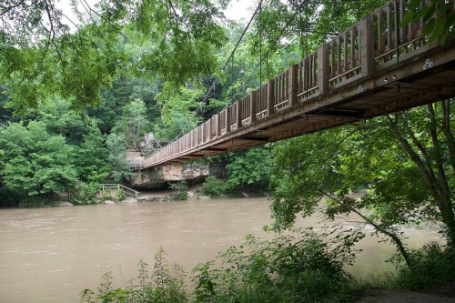A wooden suspension bridge spans a river, surrounded by lush green trees and rocky cliffs.