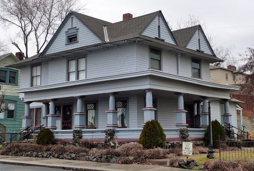 A large, two-story blue Victorian house with a wraparound porch and decorative columns, surrounded by shrubs.
