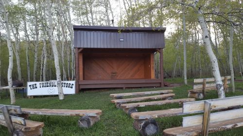 A wooden stage in a forested area, with benches arranged in front and a sign reading "The Friendly Store."