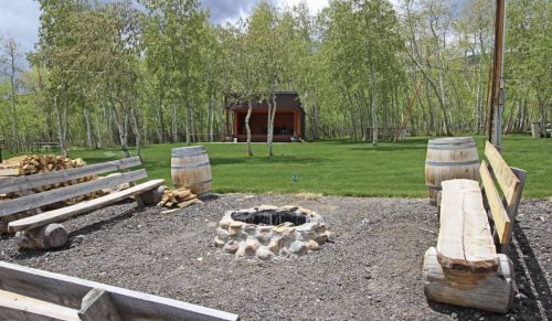 A rustic outdoor seating area with benches around a stone fire pit, surrounded by trees and a grassy lawn.