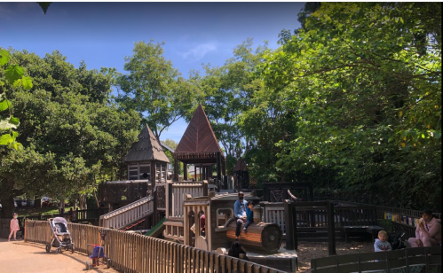 A playground with wooden structures, trees, and children playing, set under a clear blue sky.