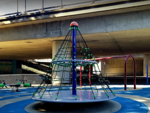 A playground features a climbing structure with a net, a spinning platform, and swings under a concrete overpass.