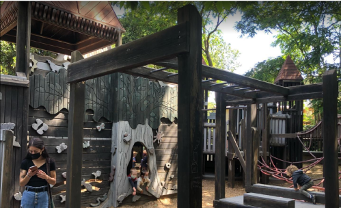 A playground with wooden structures, children playing, and an adult using a phone, surrounded by trees.