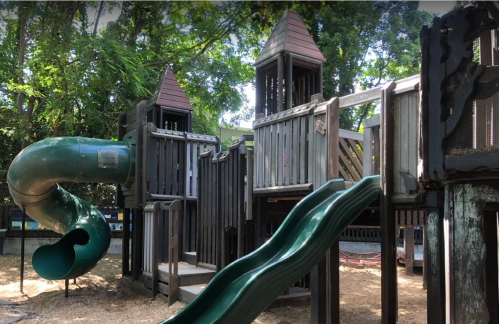 A wooden playground with two towers, a green slide, and a spiral slide surrounded by trees.
