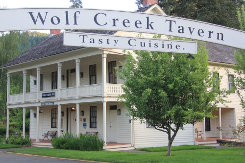 A charming white building with a sign reading "Wolf Creek Tavern" and "Tasty Cuisine" surrounded by greenery.
