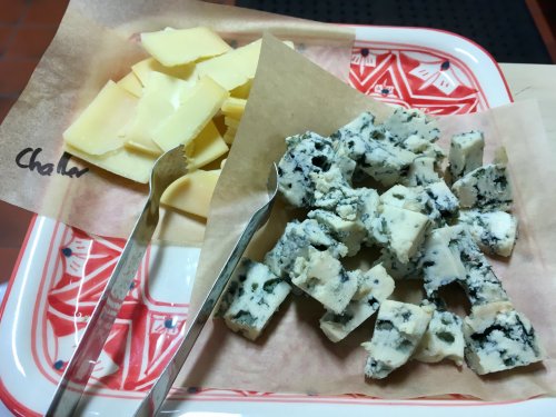 A platter with two types of cheese: yellow cheese slices on one side and blue cheese cubes on the other, with serving utensils.
