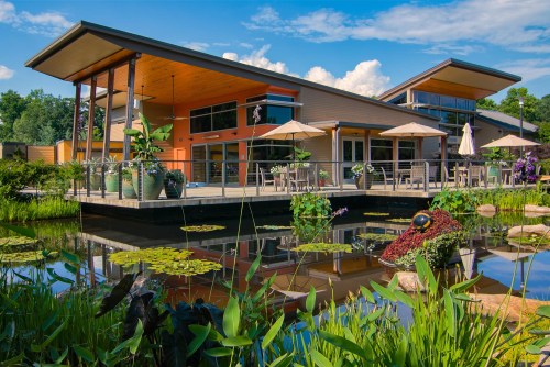 Modern building with large windows, surrounded by greenery and a pond with lily pads, under a blue sky.