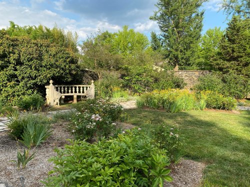 A serene garden with a stone bench, surrounded by lush greenery and colorful flowers under a partly cloudy sky.