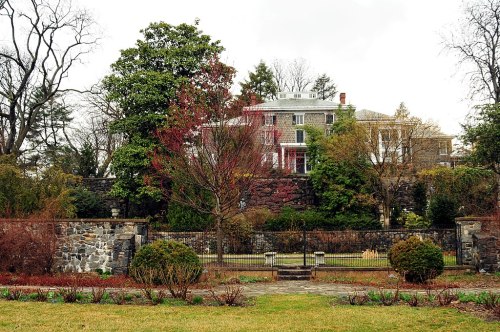 A large historic house surrounded by trees and a stone wall, with a garden in the foreground on a cloudy day.