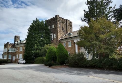 Historic stone building with a tower, surrounded by trees and a parking area under a cloudy sky.