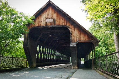 A wooden covered bridge surrounded by greenery, featuring a lattice design and a clear blue sky above.