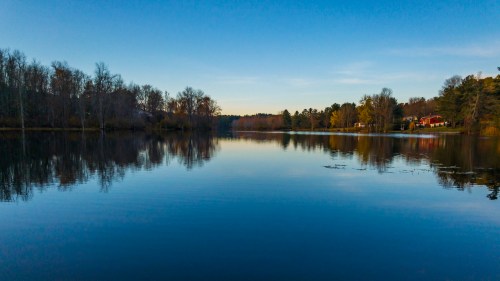 A serene lake at sunset, surrounded by trees, reflecting the sky and nearby houses on the calm water.