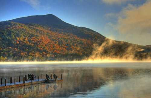 A serene lake at sunrise, surrounded by colorful autumn trees and mist rising from the water, with a dock in the foreground.